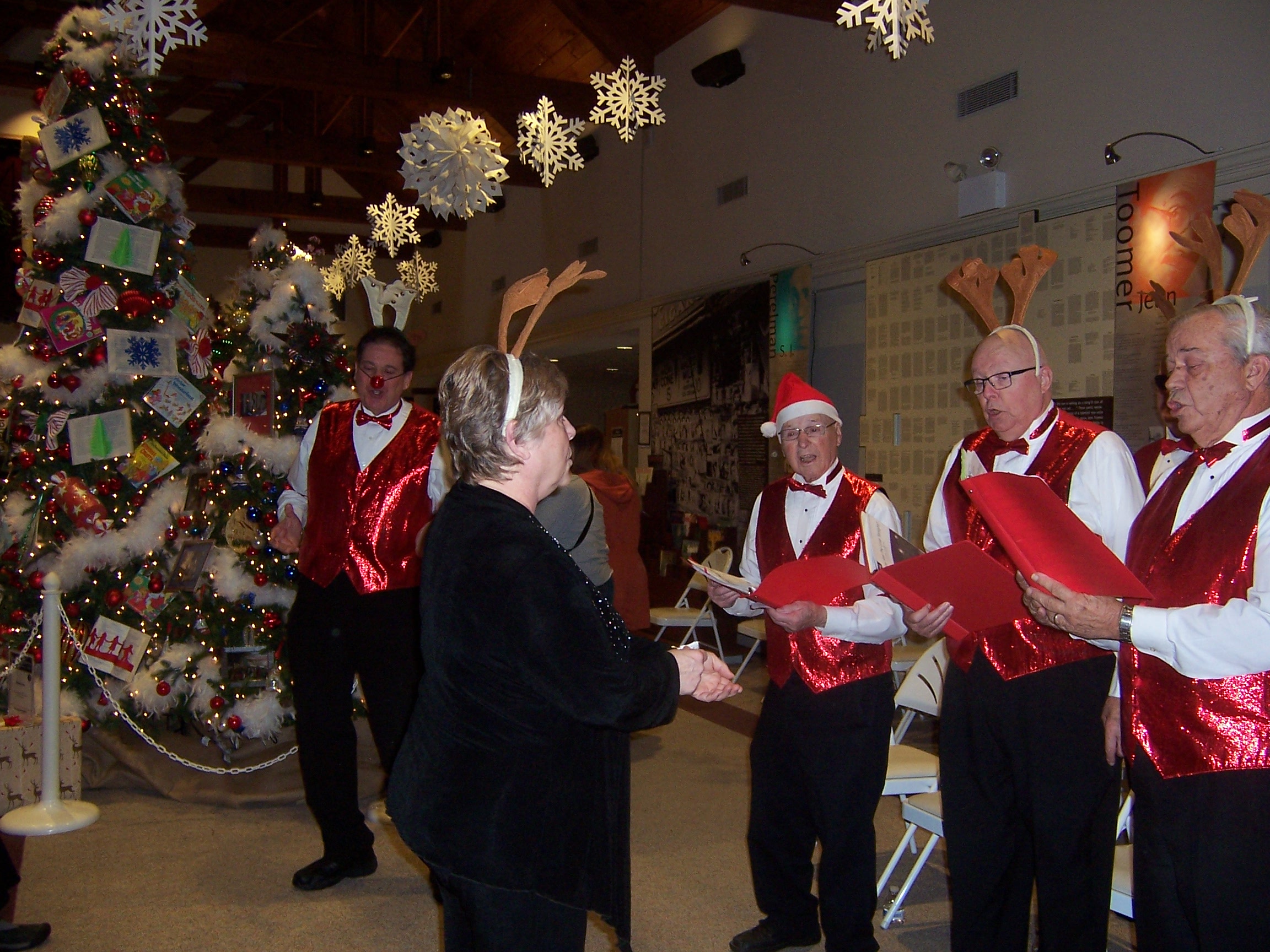 The chorus singing, onstage, in festive attire (3 of 4); Rudolph impersonator in background